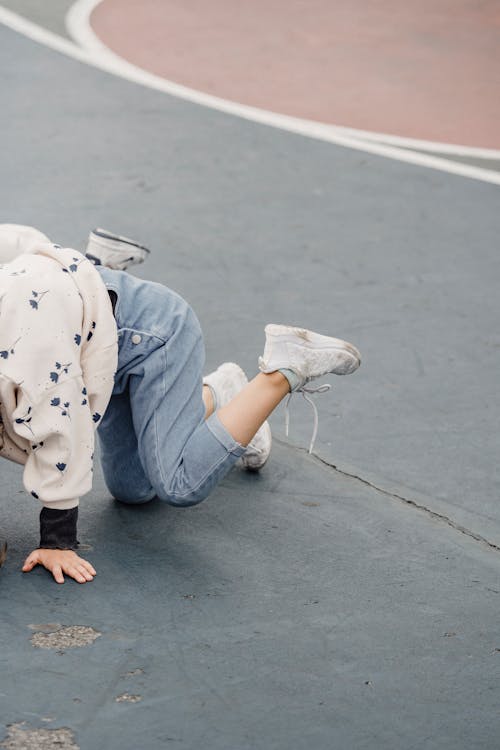Side view of anonymous playful little kid wearing jeans and sneakers standing on all fours on green shabby sports ground