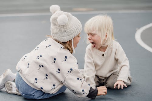 Side view of cute little girl with hat and playful boy showing tongue sitting on sports ground while having fun against blurred background