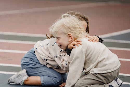 Anonymous tender girl embracing cute little boy with blond hair while sitting on colorful sports ground together  on blurred background