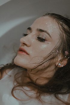 从上方看，一位身穿白衣、妆容忧郁的女性躺在浴缸的水中，目光移向别处