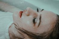 一位身穿白衣、头发湿漉漉的女士，正闭着眼睛，放松地躺在自家浴缸的水中