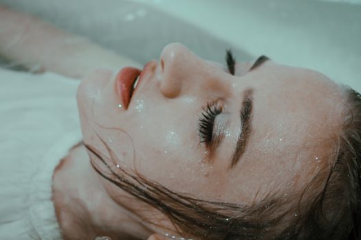 一位身穿白衣、头发湿漉漉的女士，正闭着眼睛，放松地躺在自家浴缸的水中