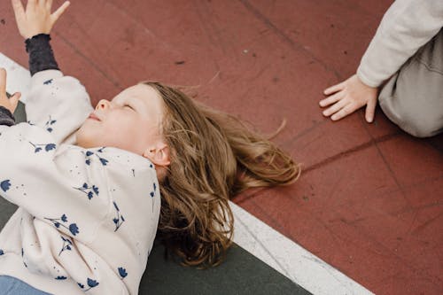 Top view of playful little girl with closed eyes lying in colorful sports ground near unrecognizable kid while having fun together