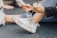Unrecognizable child with bent leg tying laces on dirty footwear while sitting on playground on street with anonymous kid on blurred background