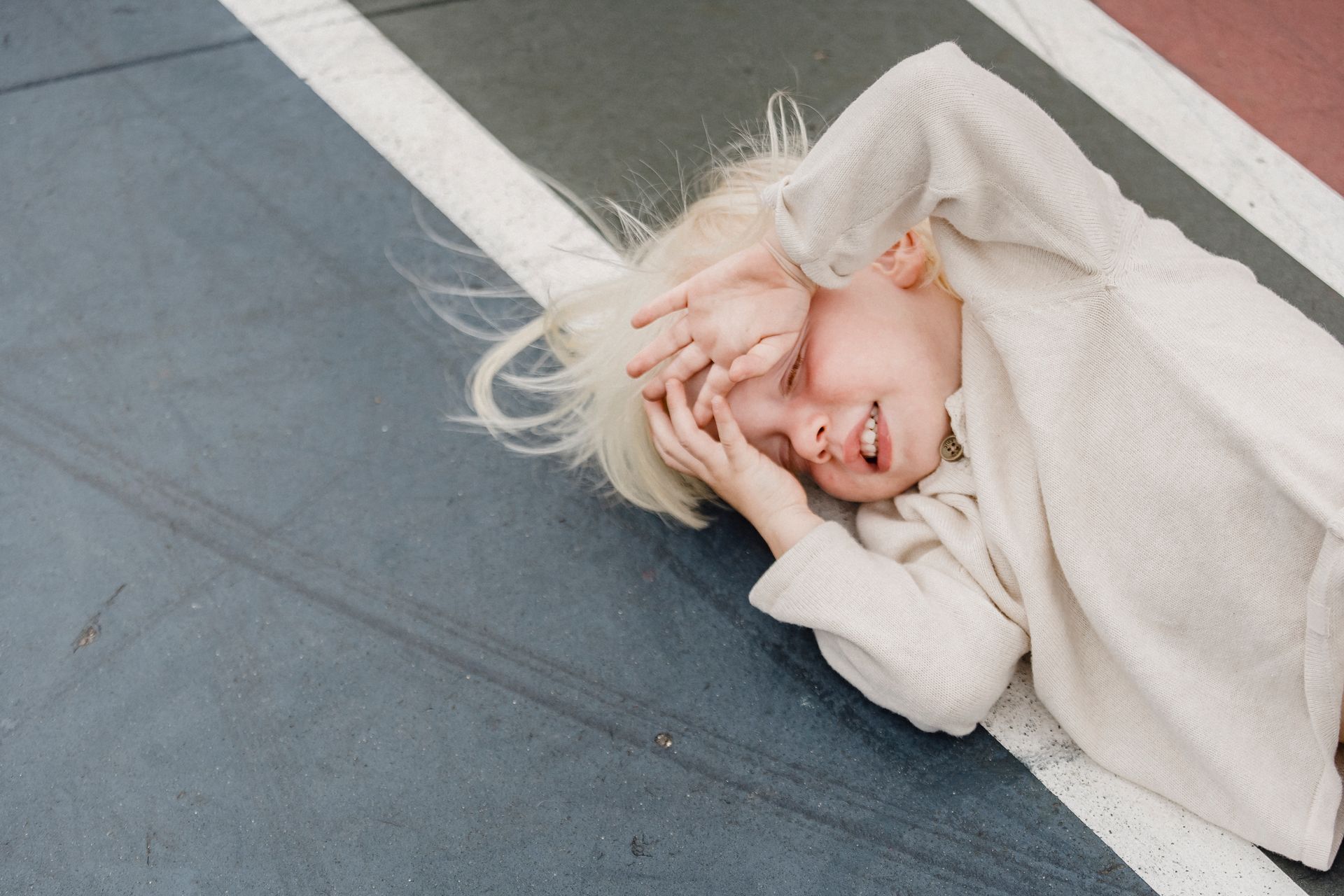From above of playful little blond kid in white shirt with closed eyes and hands on face lying on colorful sports ground