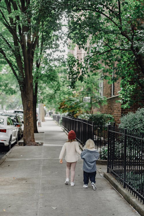 Full body back view of anonymous boy and girl strolling on pathway along metal fence and green trees on street