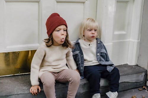 Adorable girl in hat showing tongue while sitting on step with cute little boy near entrance of building on street