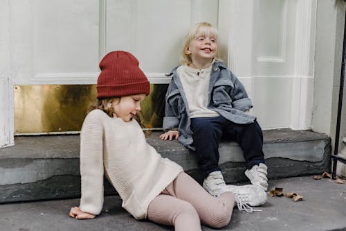 Funny girl in hair and blond boy having fun while sitting on porch near door of residential building on street