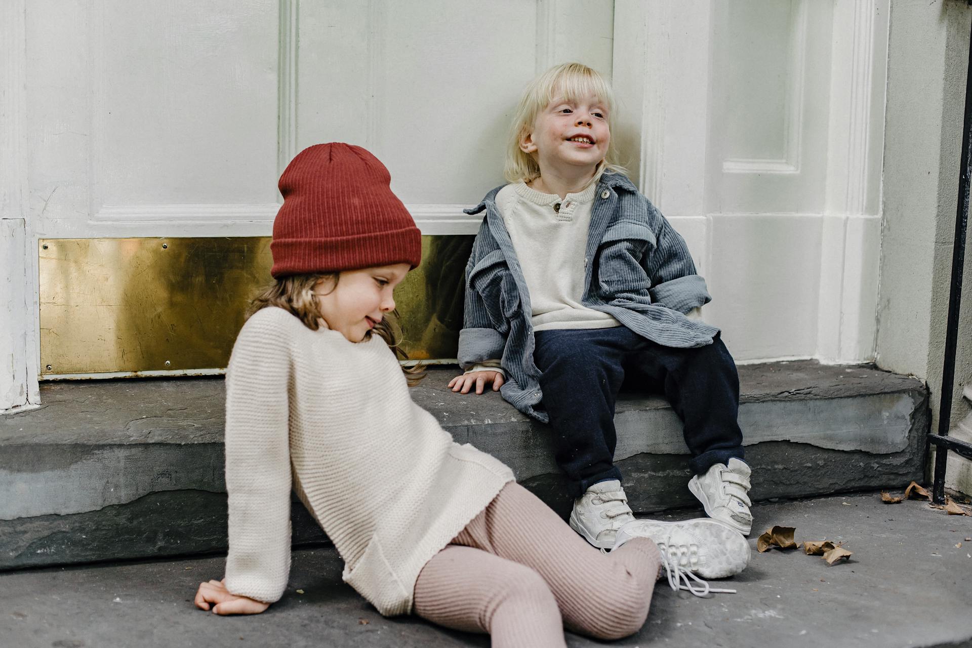 Funny girl in hair and blond boy having fun while sitting on porch near door of residential building on street