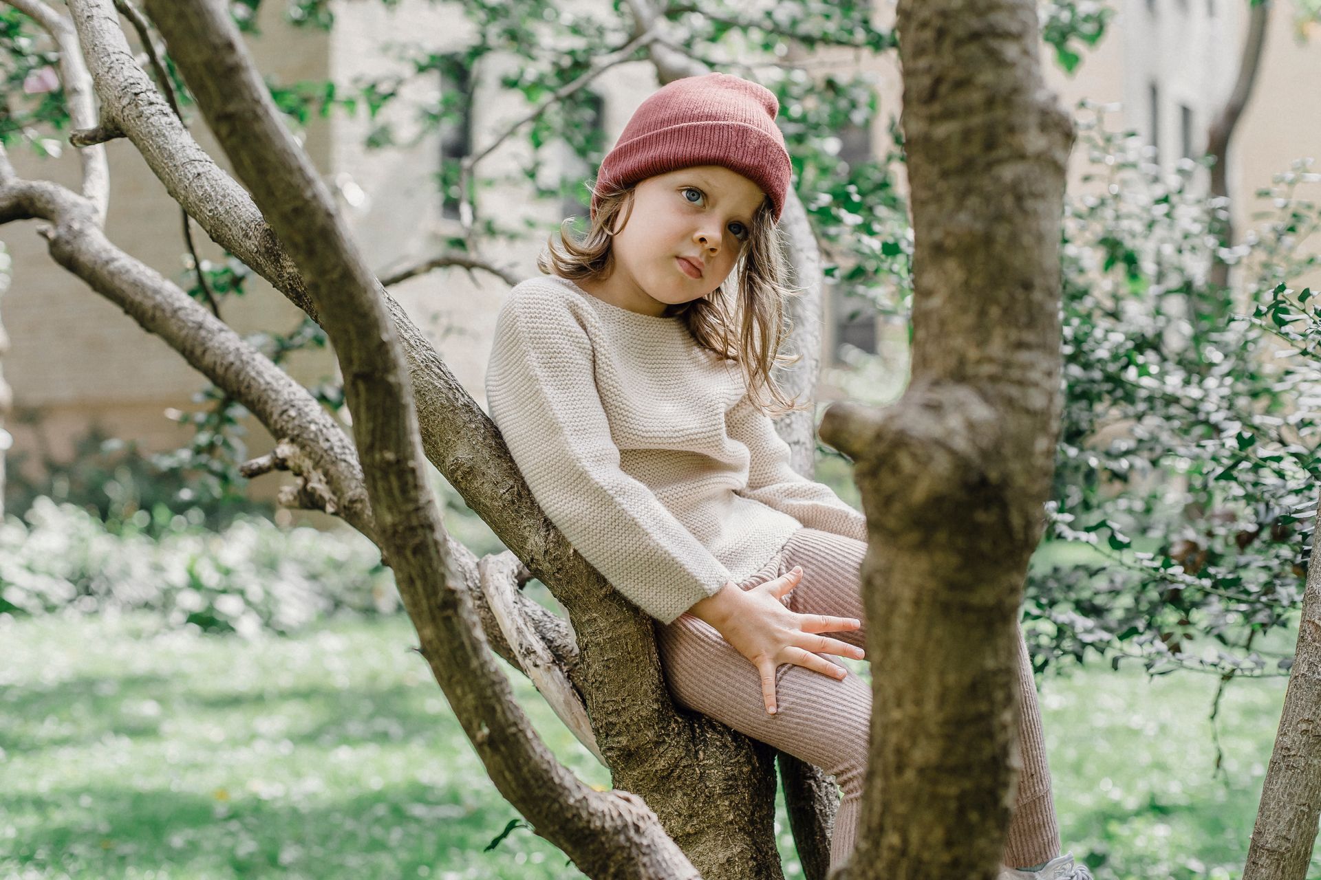 Dreamy girl wearing hat looking away while sitting on tree branch on street against building and foliage on blurred background