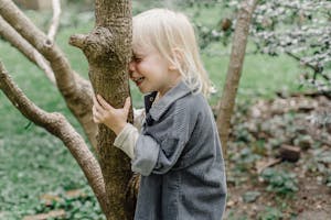 Side view of cheerful little kid with closed eyes smiling and touching tree trunk while standing in park on blurred background