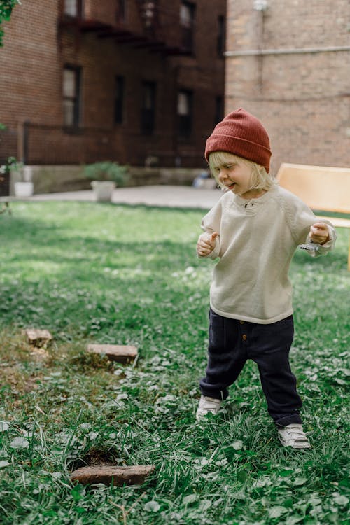 Full body of playful little kid wearing hat standing on grassy ground with toy in hand against residential building on blurred background