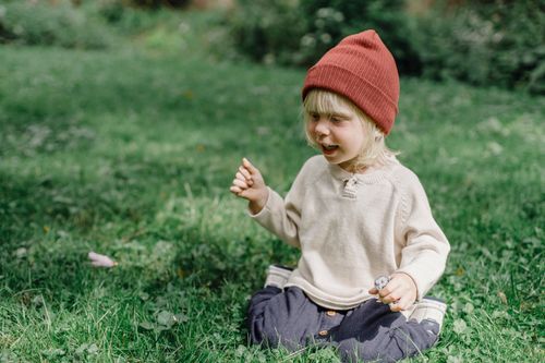 Free Full body of cheerful kid with toy in hand wearing hat smiling while sitting on grassy lawn on blurred background Stock Photo