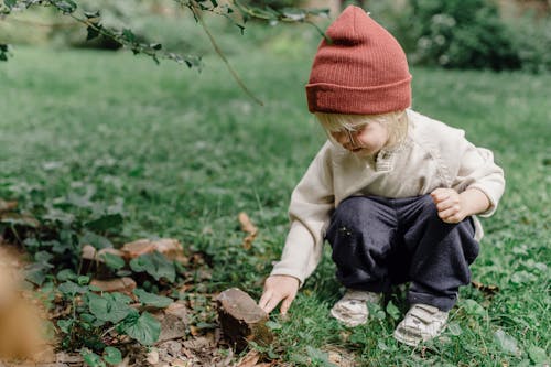 Kind In Der Orangefarbenen Strickmütze Und In Der Braunen Jacke, Die Auf Grünem Gras Sitzen