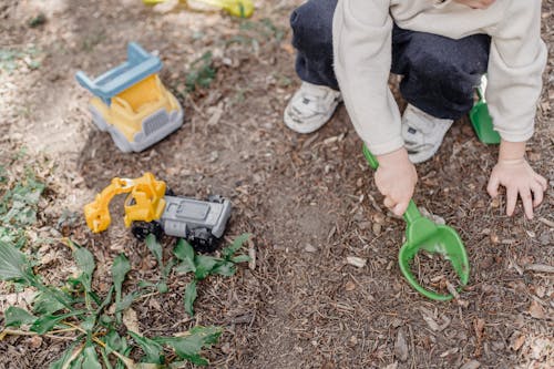 High angle of unrecognizable child playing with plastic toys sitting on ground in yard