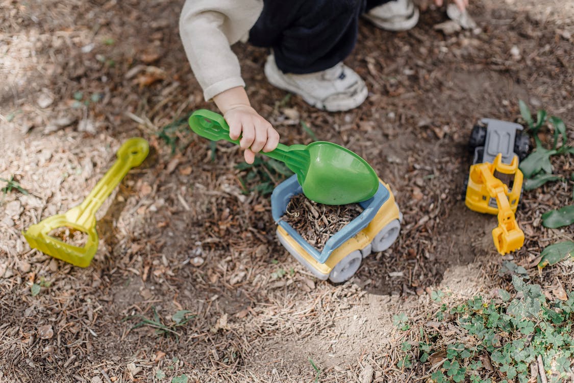 Free Unrecognizable little child playing with plastic toy car and shovel in calm sunny yard Stock Photo