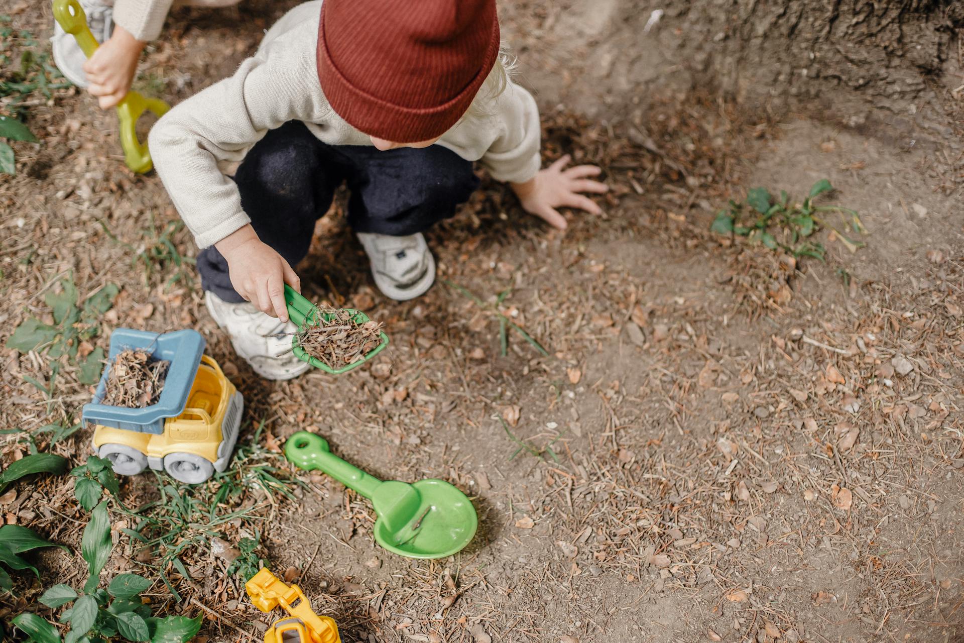 High angle of little kid in brown hat squatting on ground and playing with plastic colourful toys