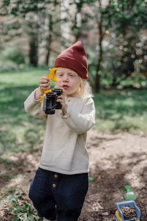 Little blond boy in casual clothes and brown hat playing with toy binoculars in garden in sunny day