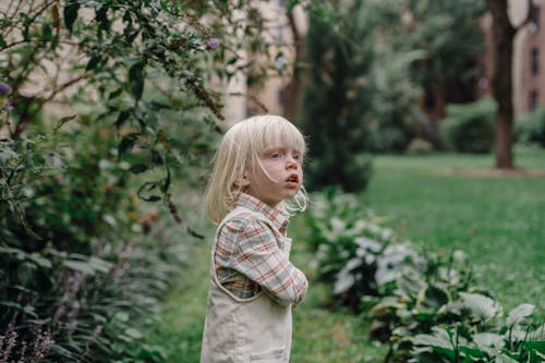 Side view of adorable little boy in casual clothes standing in green garden and looking away while spending time in garden