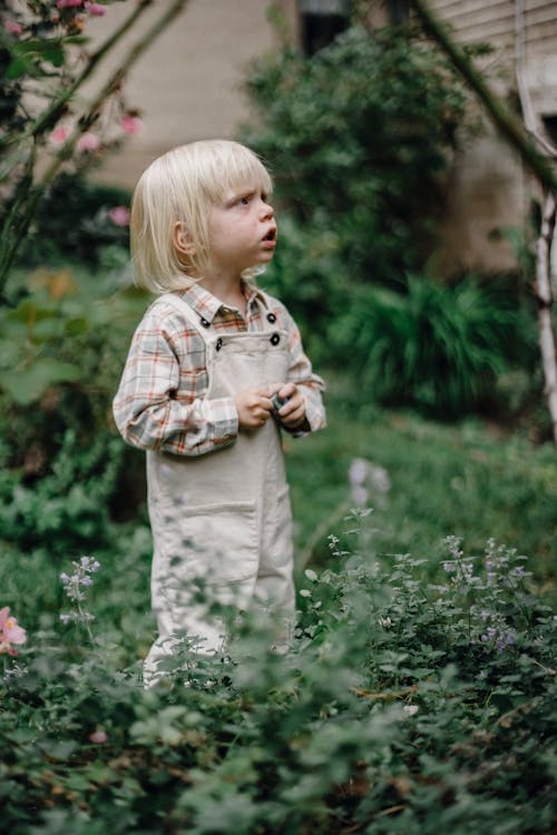 Side view of cute thoughtful little boy with blond hair in stylish clothes standing amidst lush green plants in garden and looking away