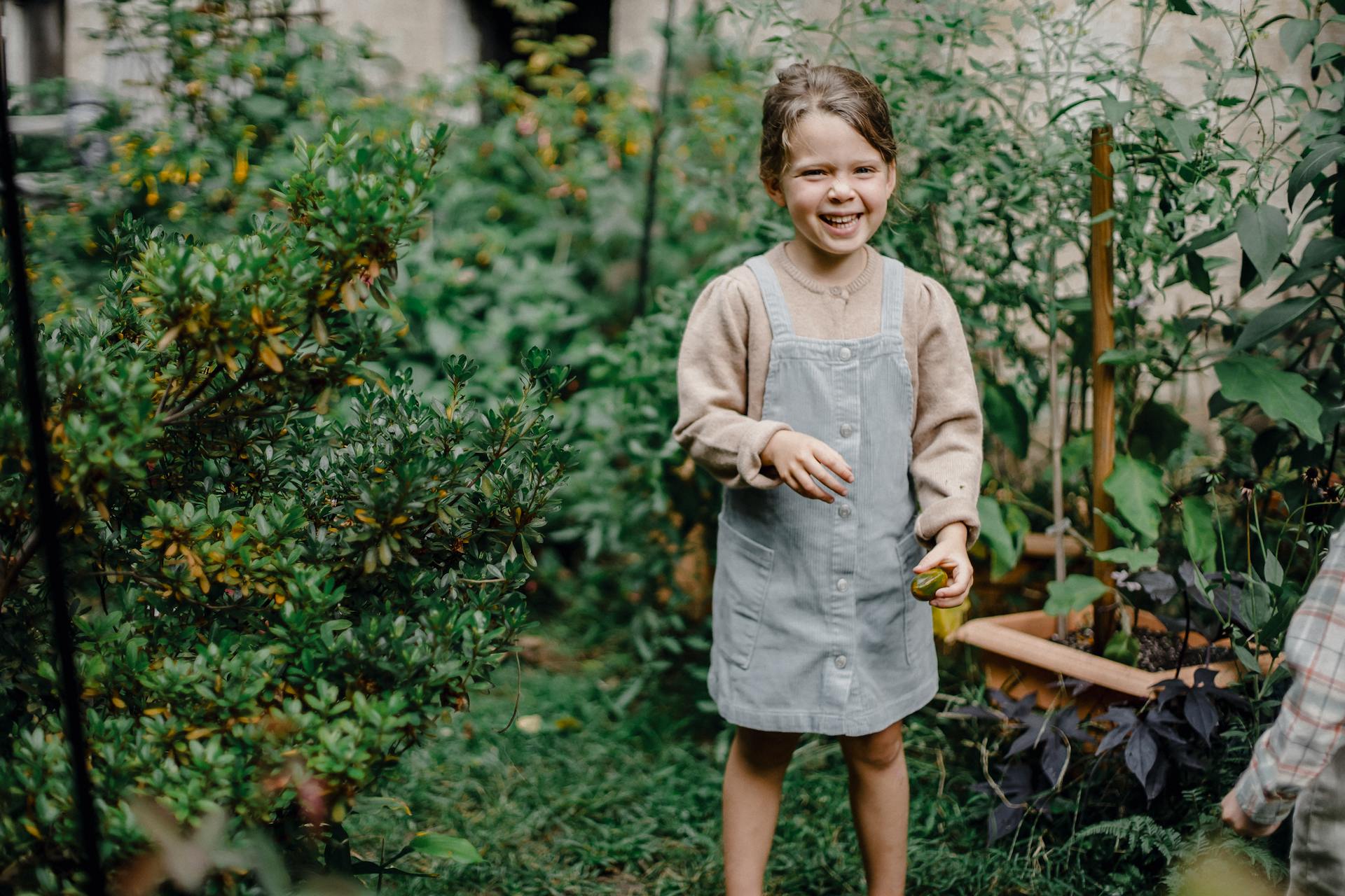 Cheerful little girl in stylish outfit smiling while standing in garden amidst lush green shrubs with picked fruit in hand