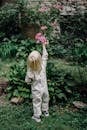 Back view of anonymous little child with blond hair in stylish clothes picking roses from shrub while resting in green park