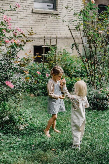 Full body of little sister and brother with blond hair in stylish clothes eating freshly picked fruits while standing on lawn in garden
