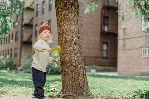 Adorable funny boy in casual warm outfit playing with plastic rake and shovel near tree and buildings in yard