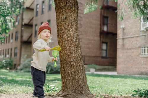 Free Adorable funny boy in casual warm outfit playing with plastic rake and shovel near tree and buildings in yard Stock Photo