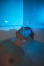 Adult man lying in bathtub full of water and looking up in dark blue light