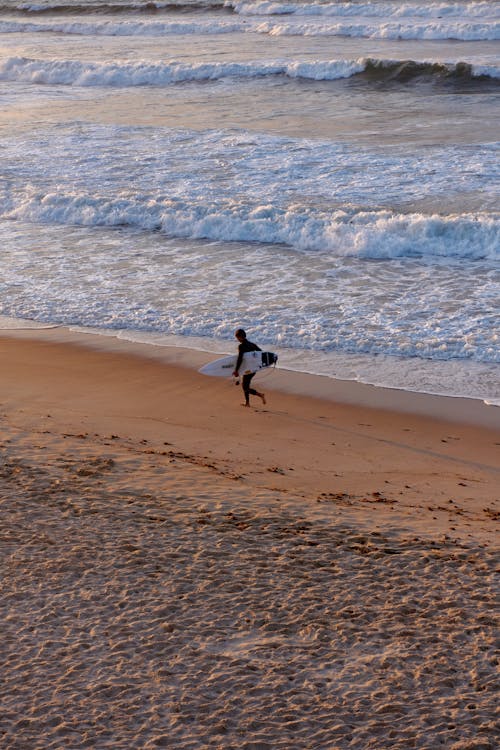 Photo of a Surfer in a Wetsuit Carrying His Surfboard while Running