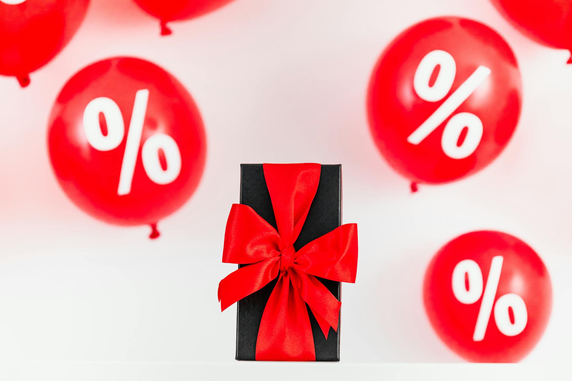 A Gift With Red Ribbon in Between Red Balloons With Percentage Symbols on a White Background