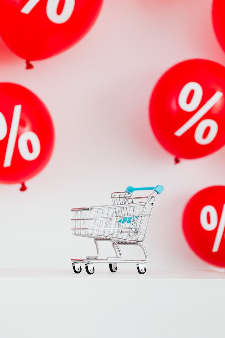 A Miniature Shopping Cart Against Red Balloons On White Background