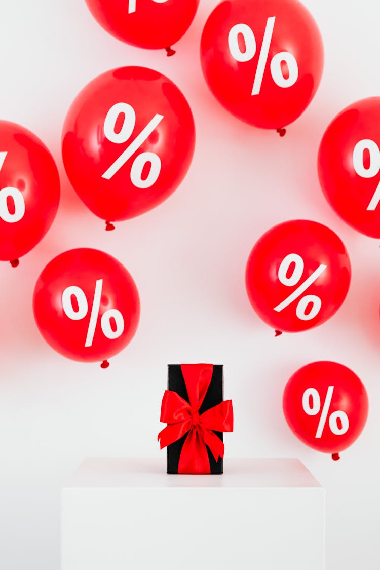 A Gift With Red Ribbon In Between Red Balloons With Percentage Symbols On A White Background