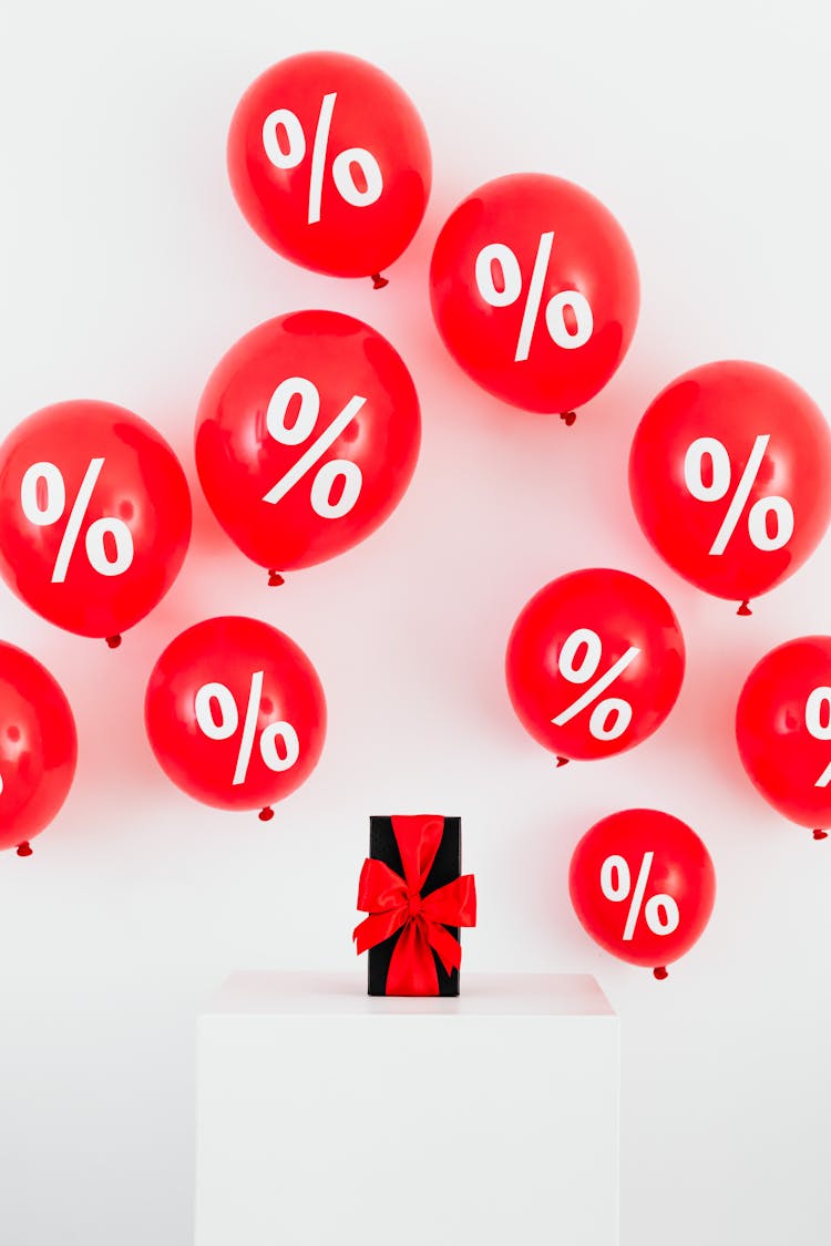 A Gift With Red Ribbon In Between Red Balloons With Percentage Symbols On A White Background