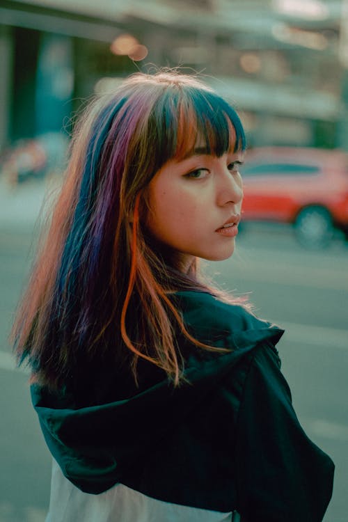 Back view of confident Asian teenage lady with colorful hair in casual outfit standing on city street and looking over shoulder thoughtfully