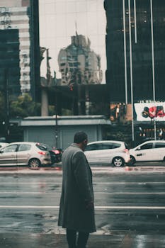 在城市场景中，一个穿着外套的男人、汽车和雨天建筑物的倒影