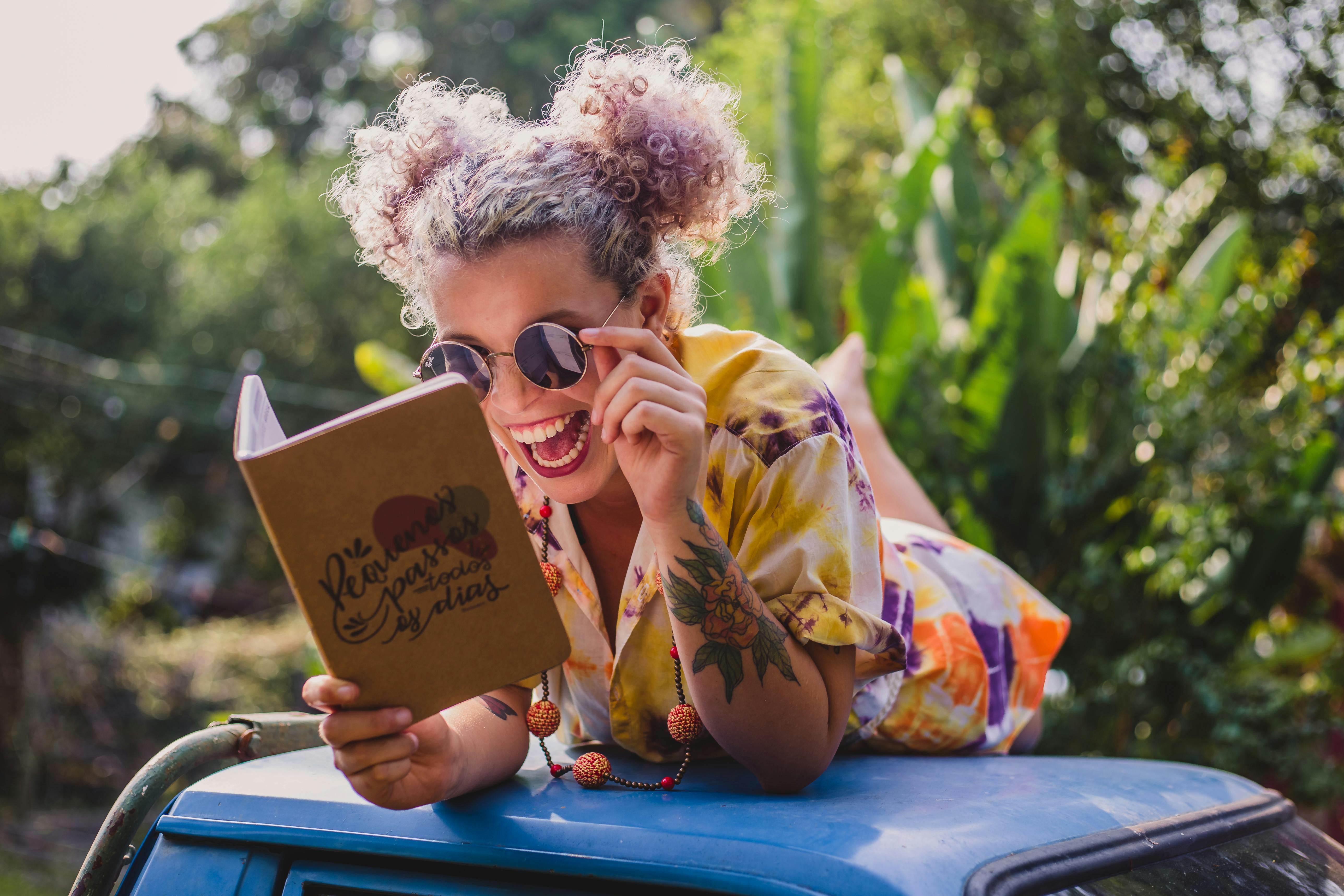 Expressive young tattooed lady with curly hair in summer outfit and sunglasses lying on car roof and reading funny book