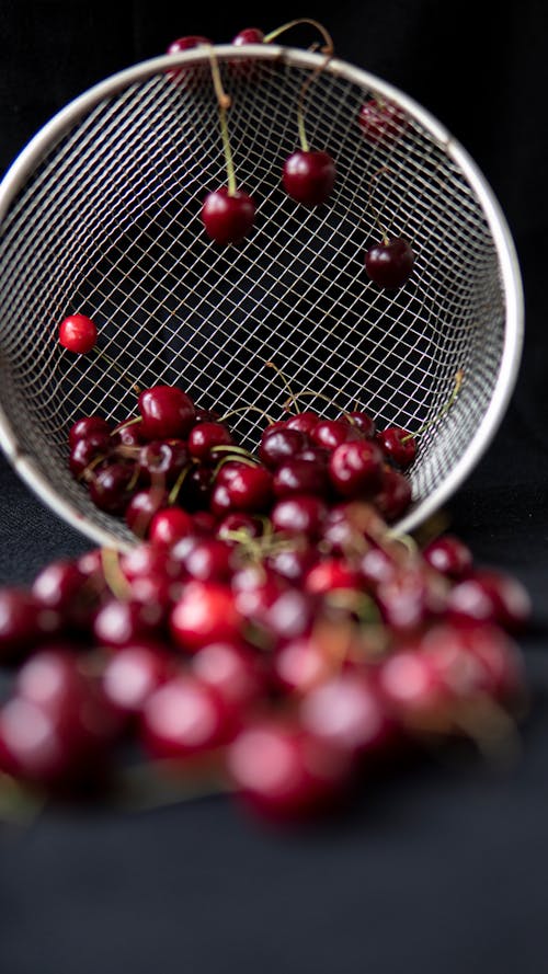 Round shaped metal strainer with pile of bright tasty cherries with shiny surface on black background