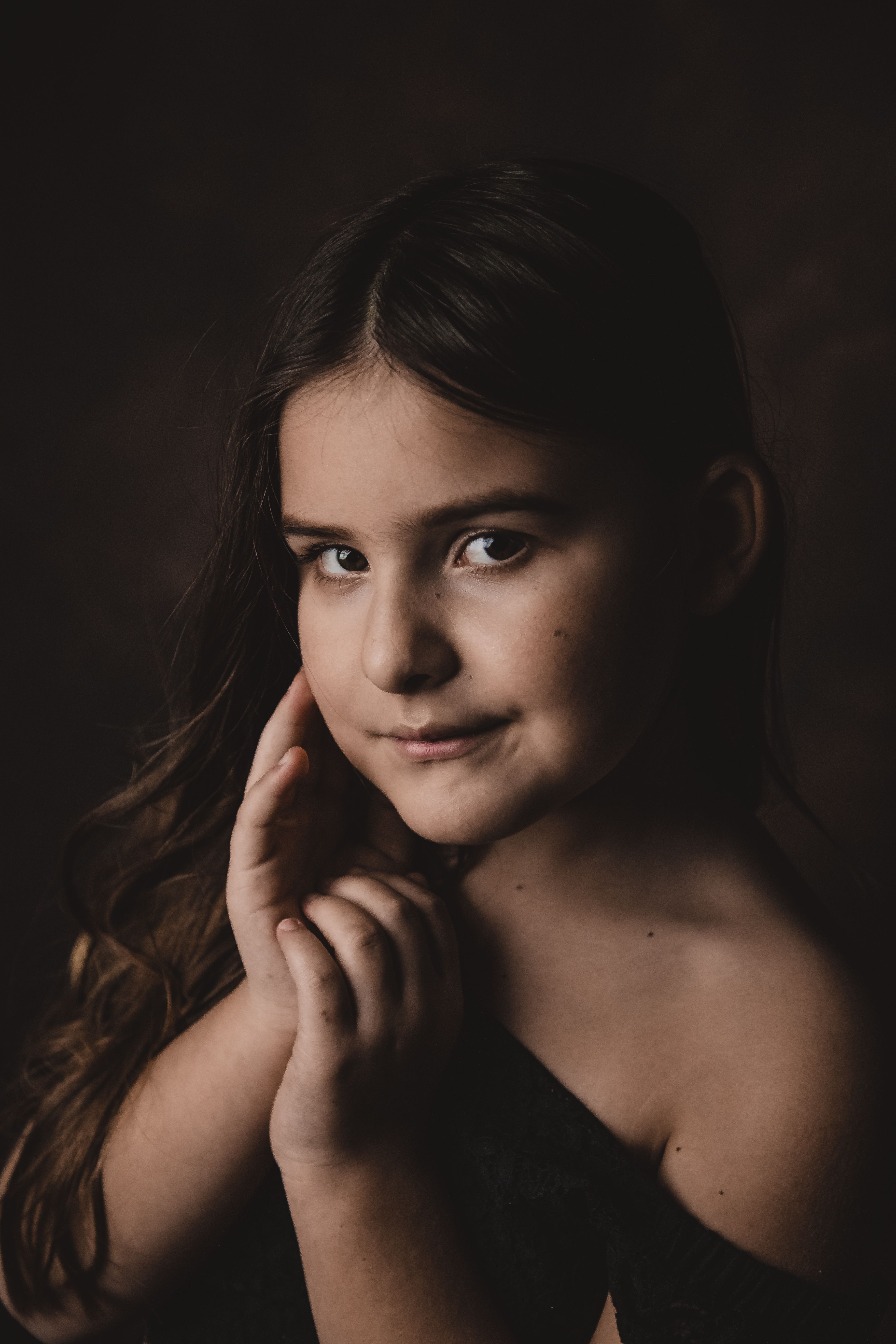 From above of attentive charming kid in trendy wear touching cheek while looking at camera