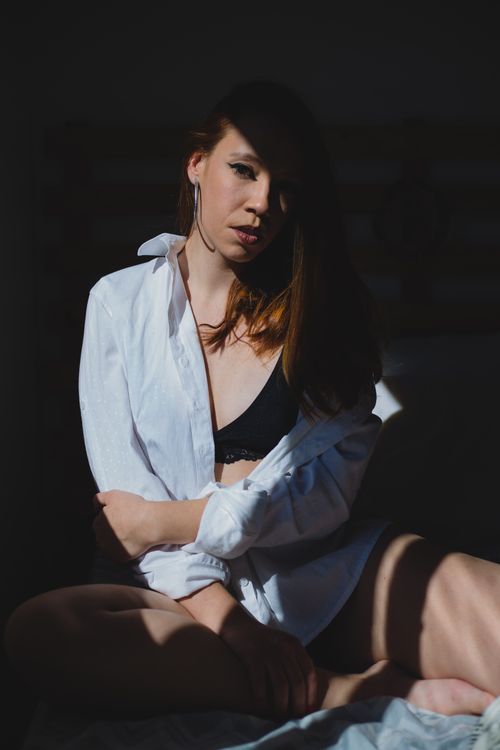 Free Young concentrated seductive female with hair on face looking at camera while sitting on bed on black background Stock Photo