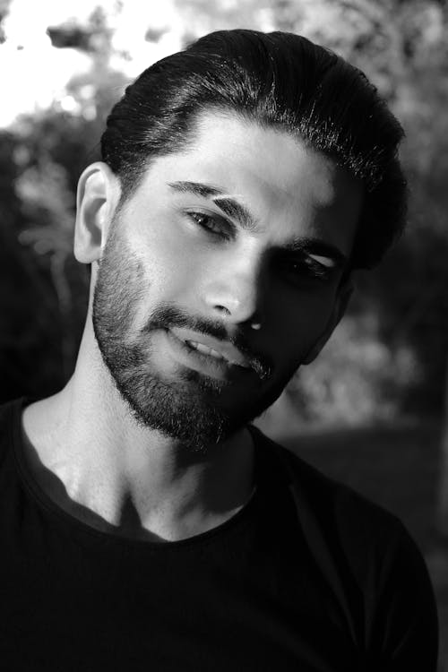 Black and white portrait of charismatic unshaven ethnic man in casual summer outfit with slicked back hair and scar on eyebrow looking at camera