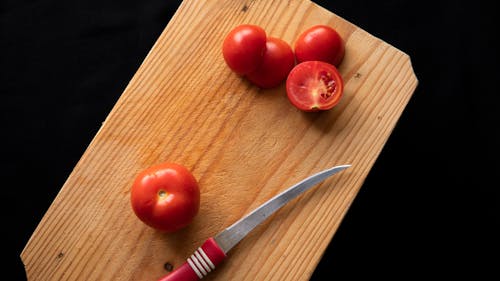Top view of thin sharp knife placed near red fresh tomatoes on timber board on black background