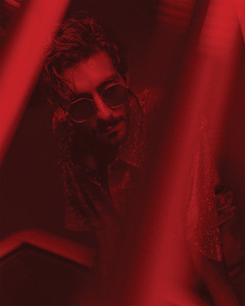 High angle of handsome male with beard and hairstyle in sunglasses standing under red illumination