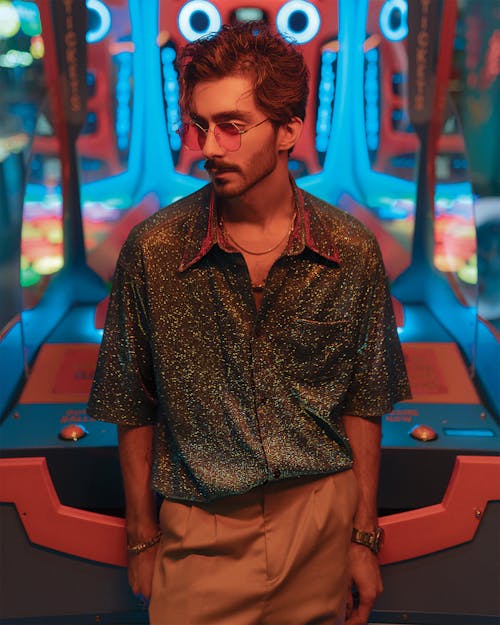 Young handsome male in trendy outfit and accessories standing near game machine in neon illumination and looking away
