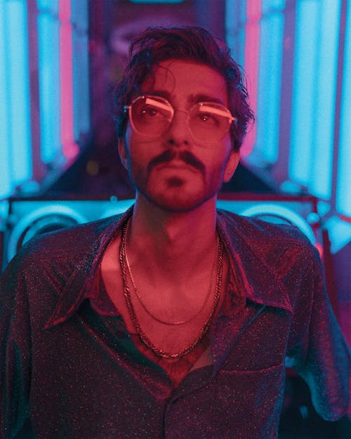Confident bearded male with glasses and chains on neck in trendy shirt looking up in room with neon lights