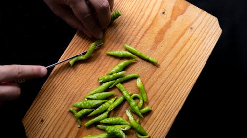 Top view of crop anonymous person cutting fresh green chili peppers on wooden chopping board