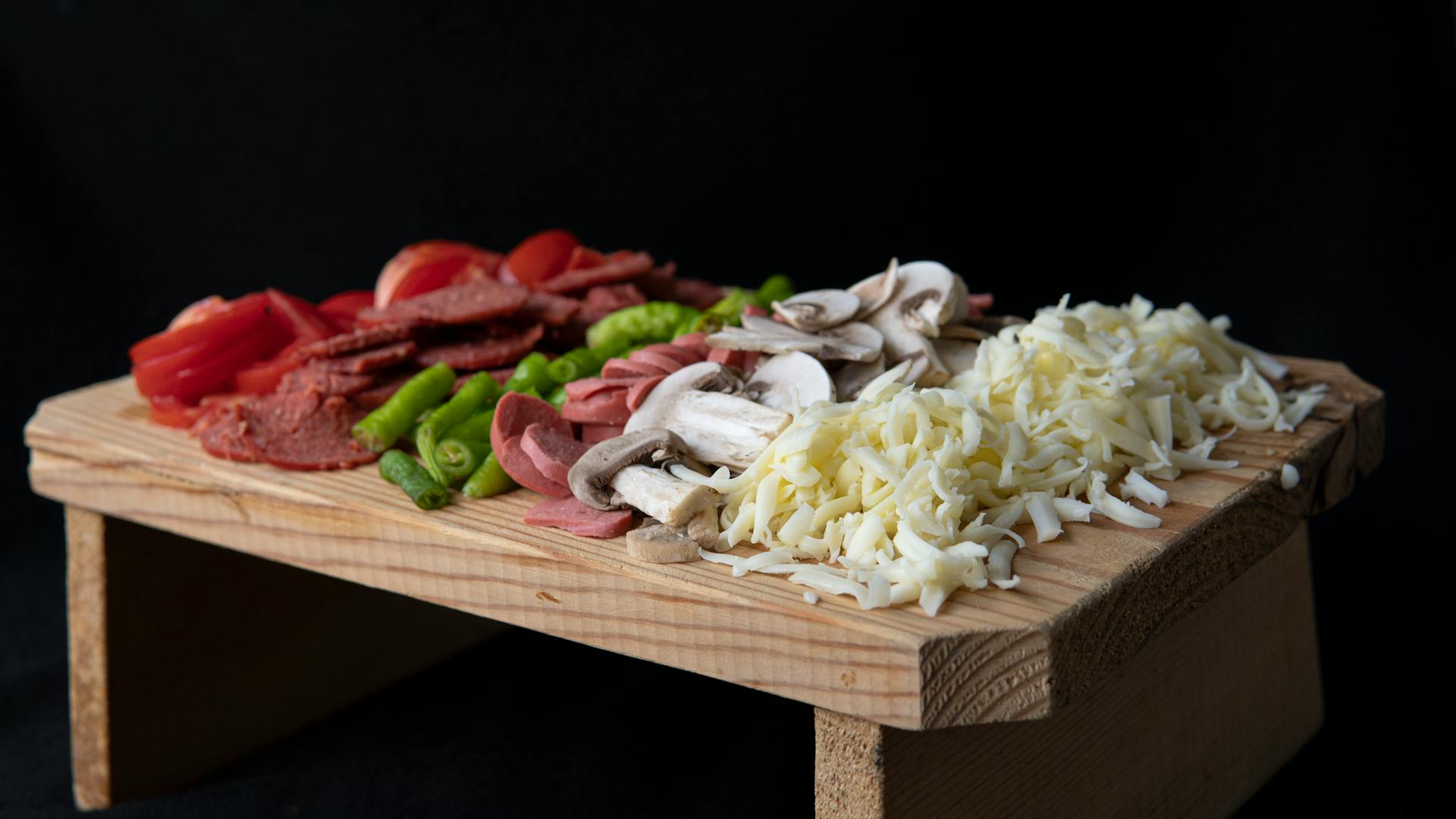 Soft focus of sliced tomatoes and hot peppers placed on chopping board with sausages and grated cheese on black background