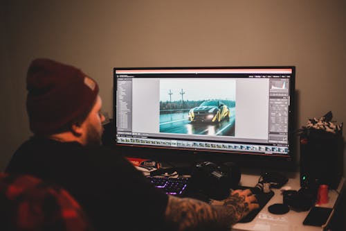 Unrecognizable tattooed male sitting at table and working on computer with montage video of sport car