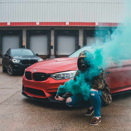 Full body of anonymous male wearing hood and respirator squatting near red car with green smoke flare in hand on street